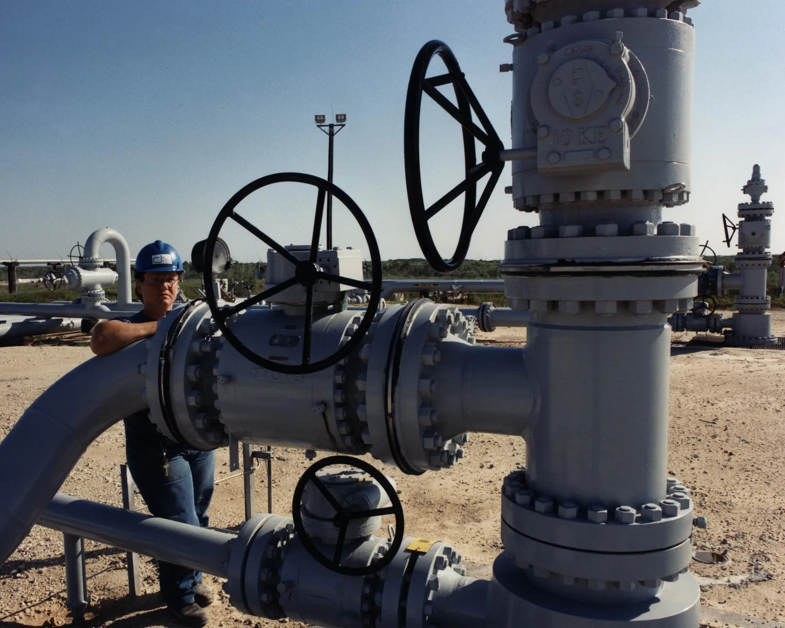 Baretli bir teknisyen, büyük vana çarkları olan gri bir petrol kuyu başı ve boru hattının yanında duruyor