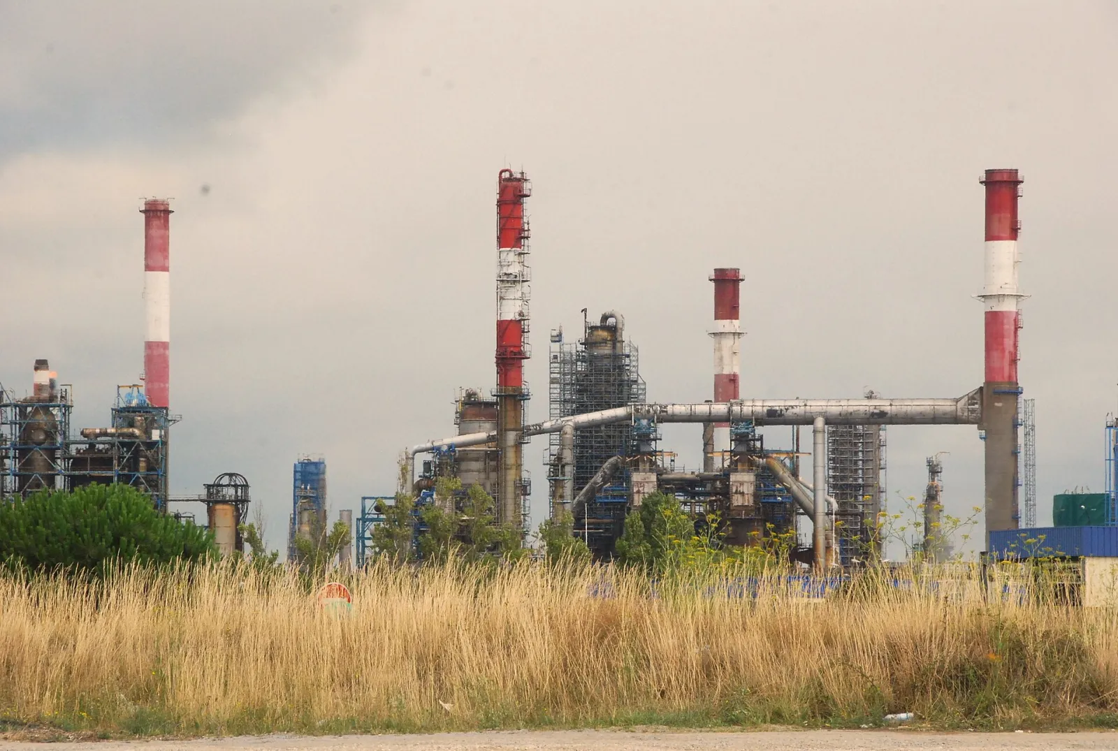 A refinery with red-and-white stacks, distillation columns and pipe racks behind dry grassland under a cloudy sky