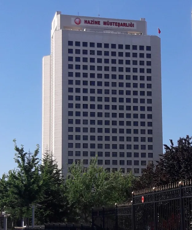 Mavi gökyüzü önünde, tepesinde HAZİNE MÜSTEŞARLIĞI yazan beyaz, çok katlı kule bina ve önünde ağaçlar