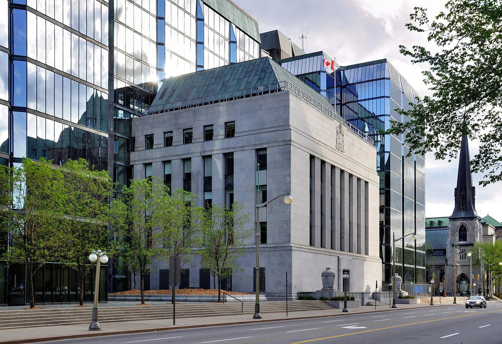 The stone-faced Bank of Canada building between glass towers in Ottawa, with the street in front