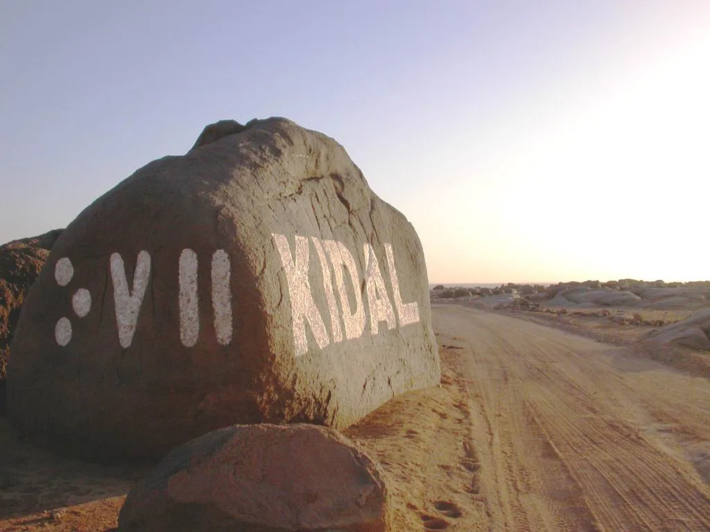 A large boulder beside a desert track with KIDAL painted on it in Tifinagh and Latin letters