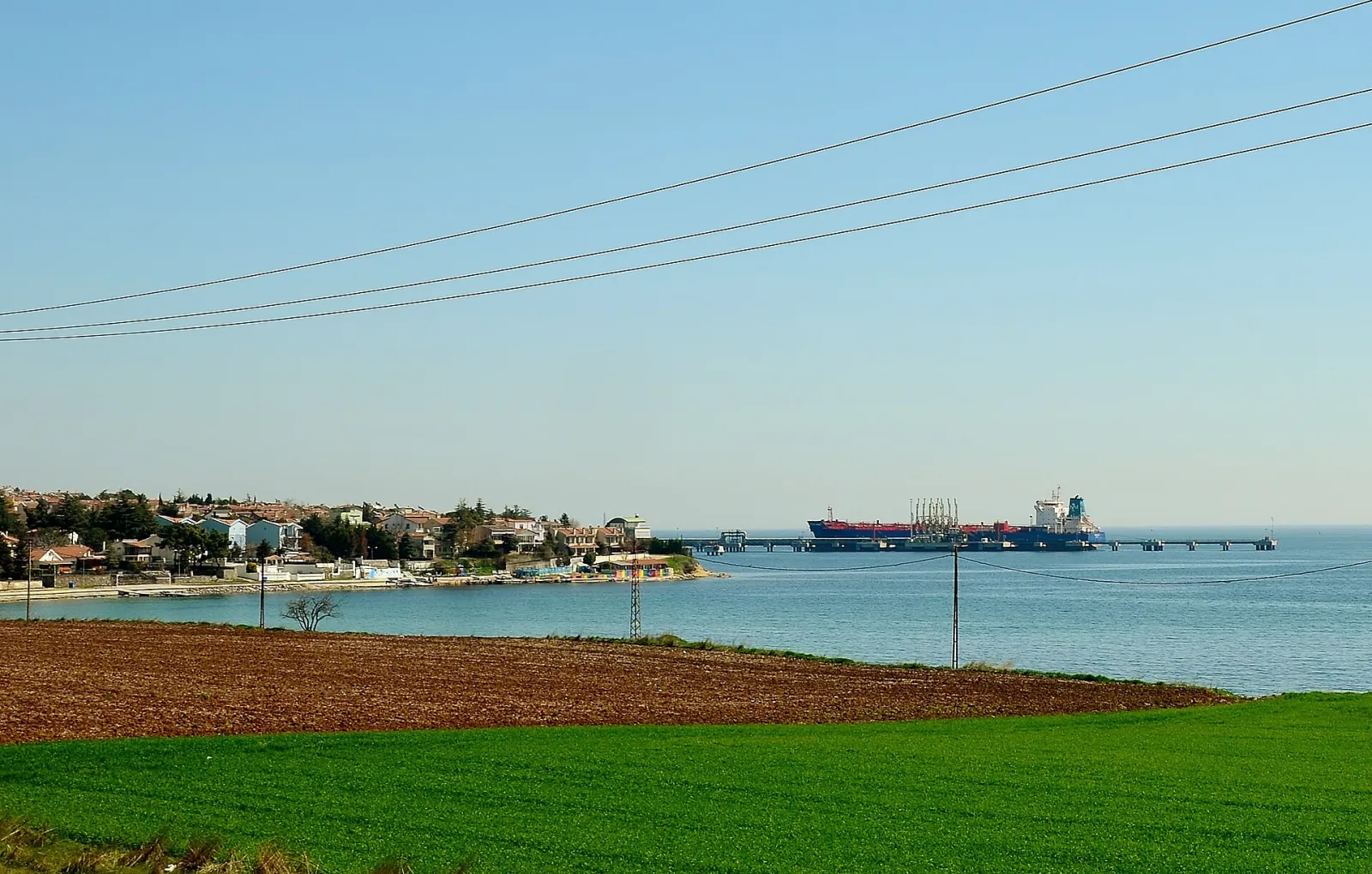 A tanker moored at the jetty of the Marmara Ereğlisi LNG storage facility, with fields and a coastal settlement in the foreground