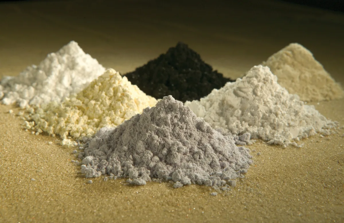Six small piles of differently coloured rare-earth oxide powders on a white surface