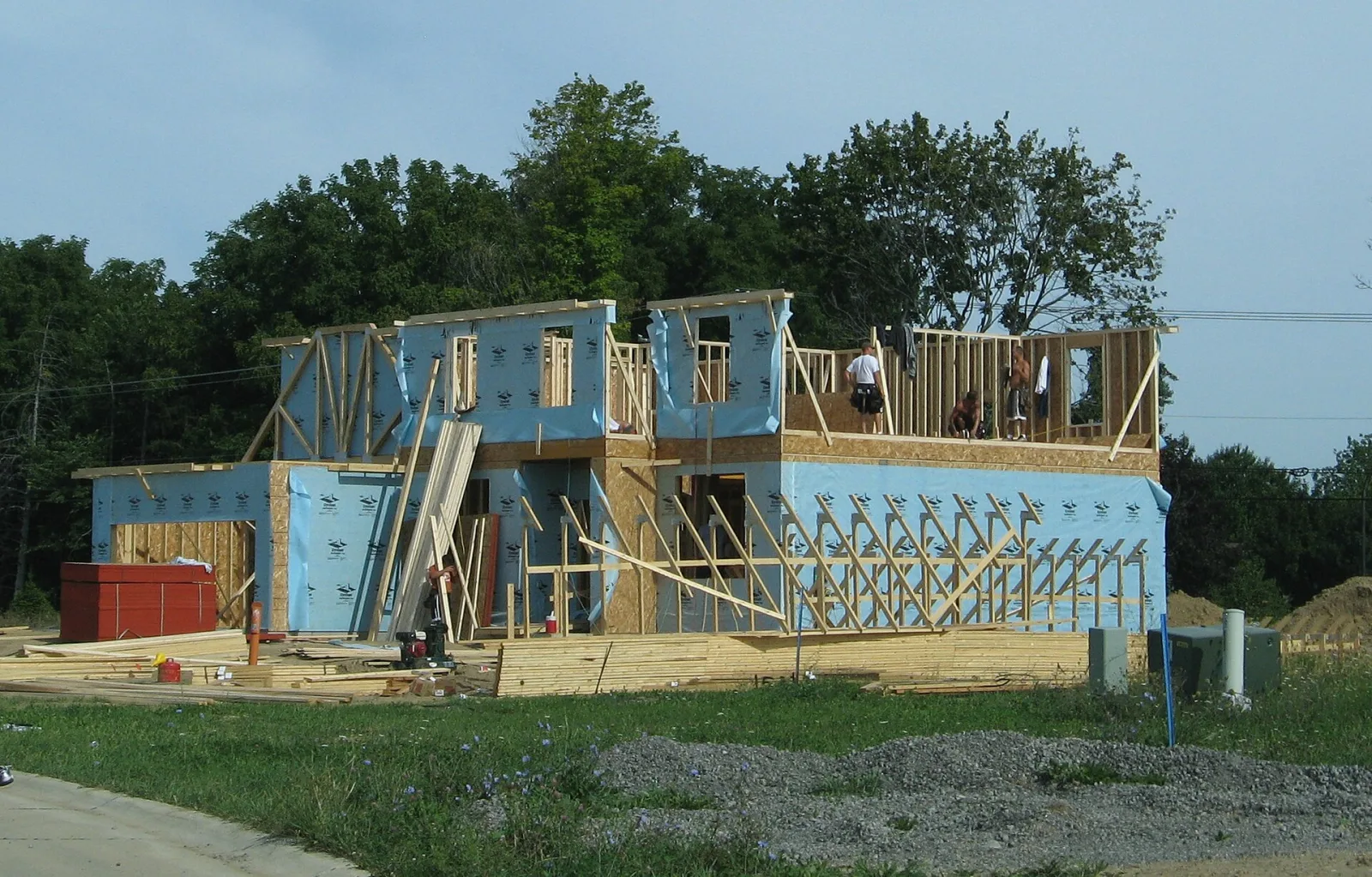 A two-storey detached house under construction with timber framing and blue house wrap, workers on the upper floor