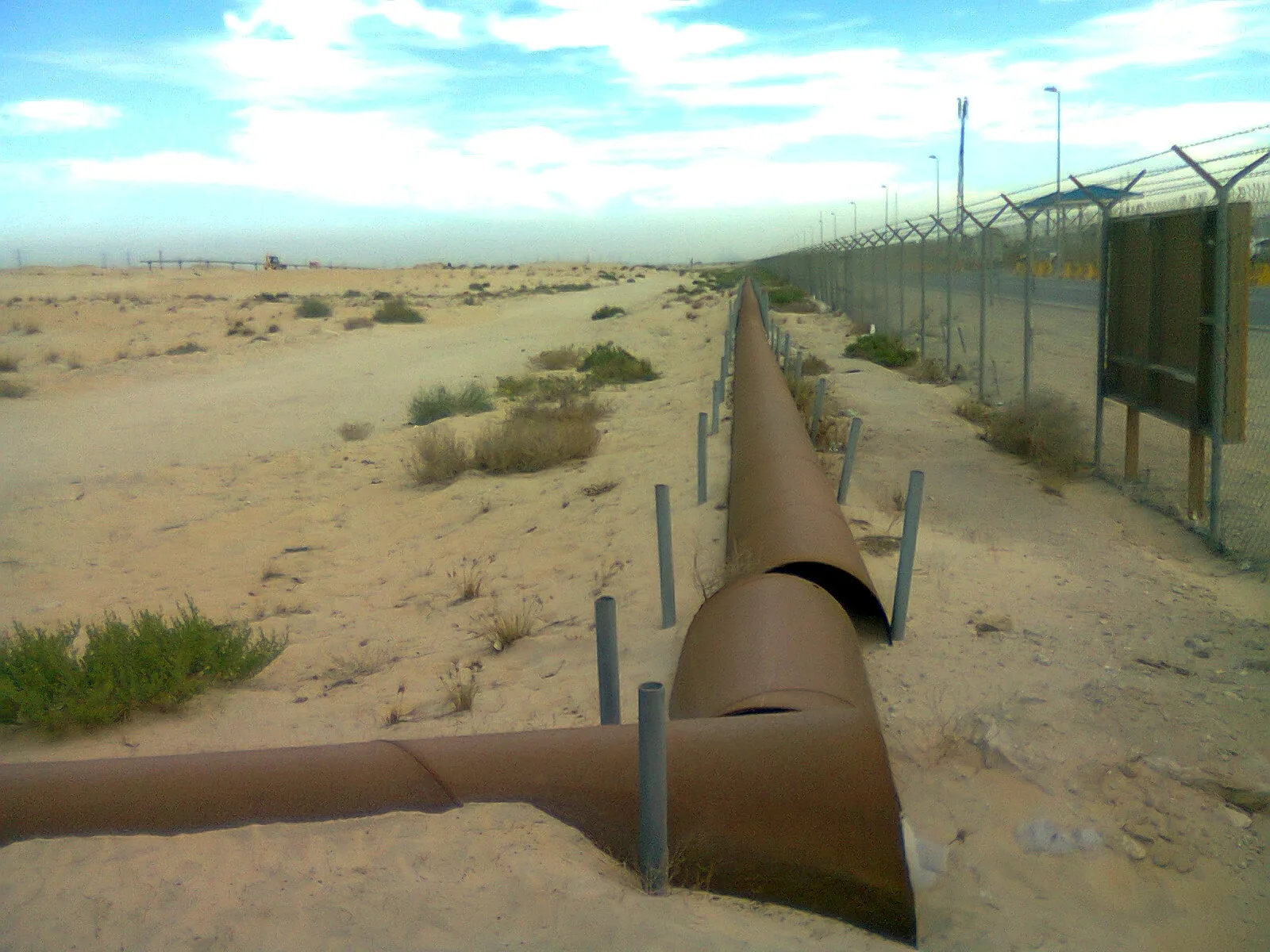 Rusty oil pipelines, partly buried, running along a wire fence through sand and scrub