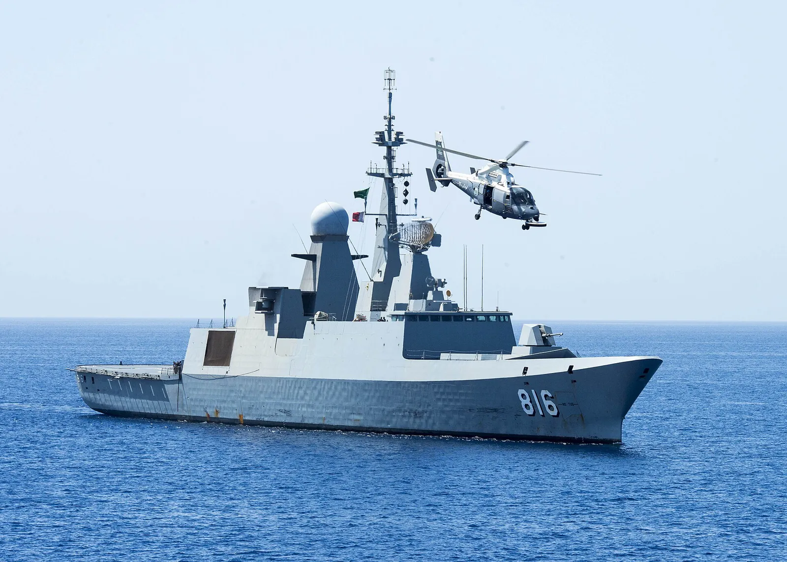 The grey Saudi frigate 816 Al Dammam under way at sea with its ship-borne helicopter circling overhead